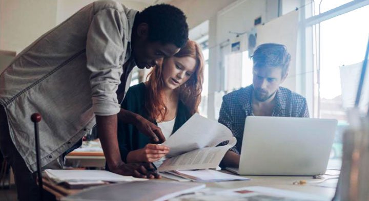 Foto de dois homens e uma mulher ao redor de uma mesa e em frente a um notebook analisando papéis.