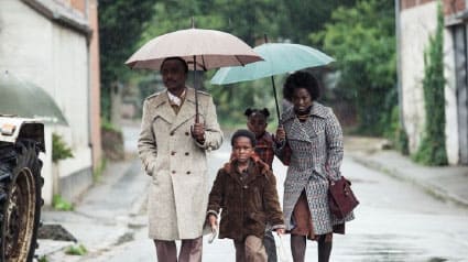 Cena do filme "Bem vindo a Marly Gomont". Casal de negros com o seu filho pequeno andando por uma rua com guarda-chuva.