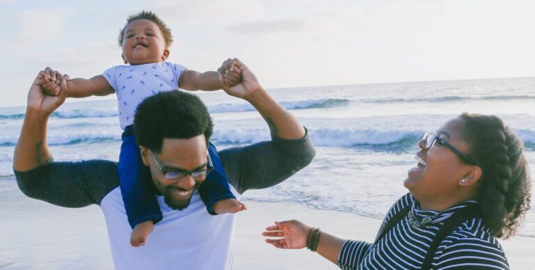 Foto de homem e mulher negros com filho de poucas semanas na praia. O bebê está sentado no cangote do pai, que o segura com as duas mãos. Todos sorriem. Licença Paternidade