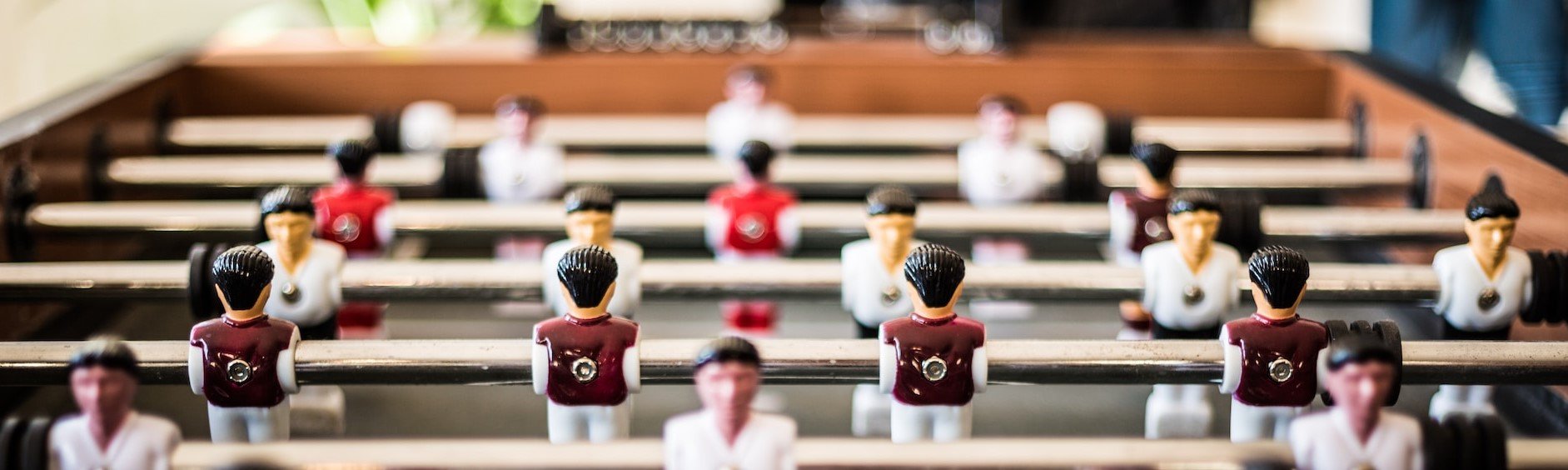 Foto em zoom de mesa de totó (pebolim) com foco nos bonecos de metal pintados e representando jogadores de futebol, 3 por linha presos uma barra de metal. A imagem busca representar a gestão desumanizada em contextos de alta pressão.