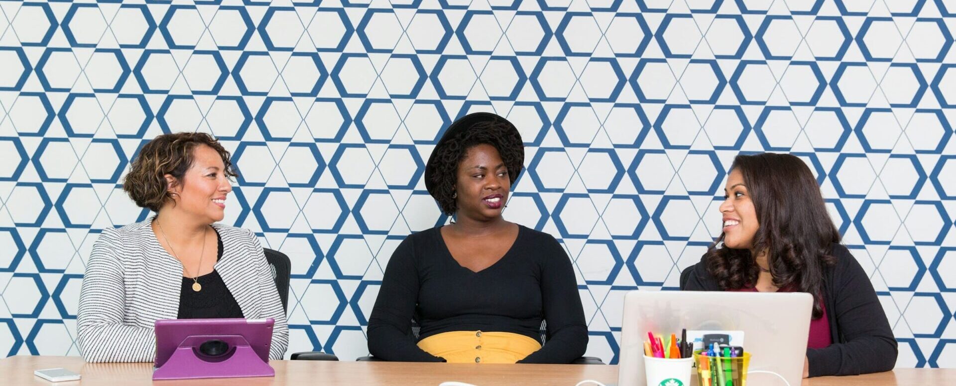 Foto de uma mulher latina, uma negra e outra caucasiana, sentadas em uma grande mesa de escritório em fundo de parede em mosaico azul e branco. Dia Internacional da Mulher #8M